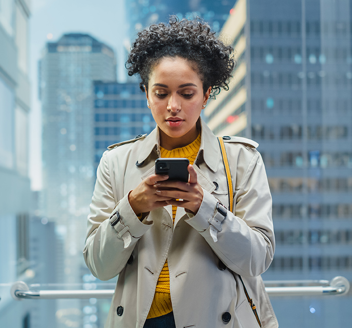 A photo of a woman in a doorway looking at a smart phone, with tall city skyscrapers behind her
