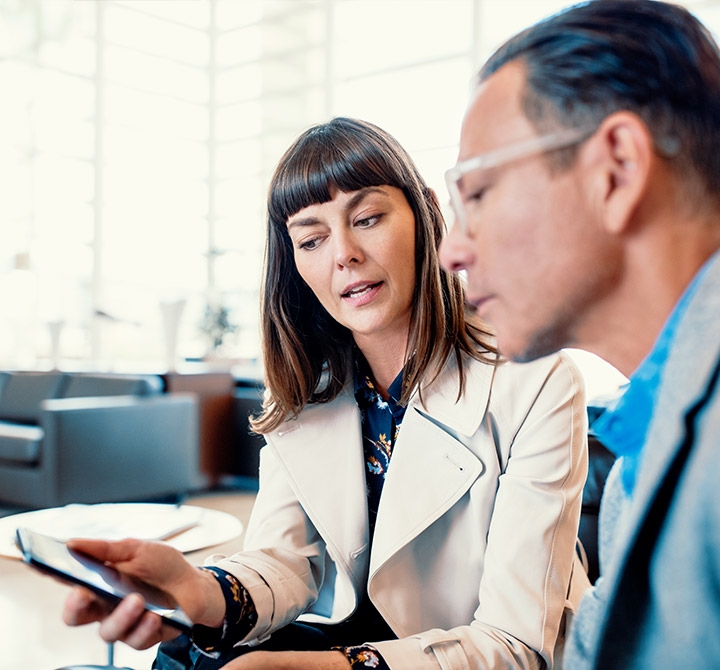 Corporate women in light-grey suite showing her mobile phone in a hand to a business man in a business meeting.