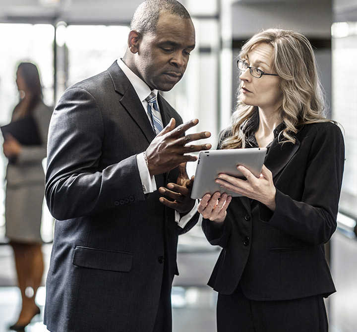 Man and woman in office setting talking while sharing a tablet.