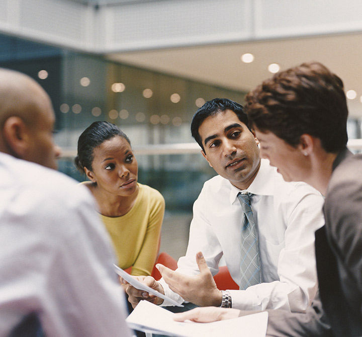 Group of four adults in an office setting holding papers and talking. 