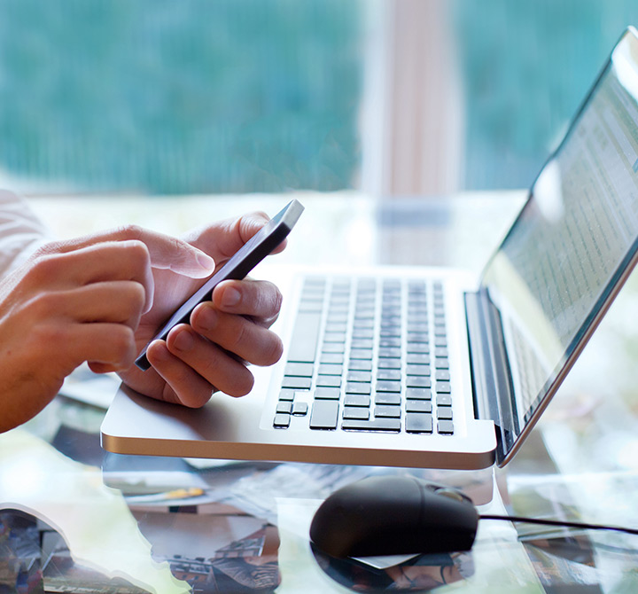 Hands holding a smartphone with a laptop and mouse sitting on a table top. 