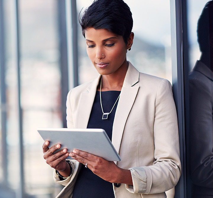 A business woman leaning against a window looking at a tablet.