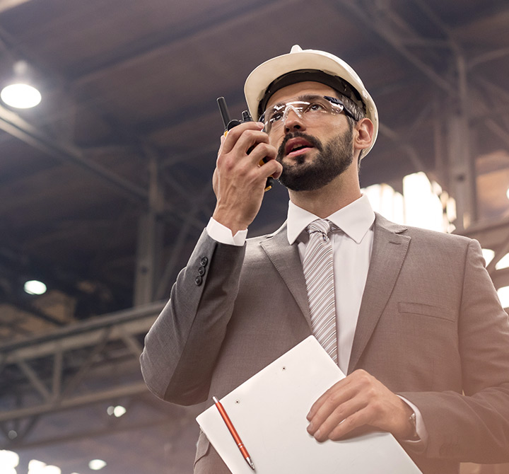 Man in warehouse on a two way radio wearing a suit, hardhat, and carrying a clipboard.