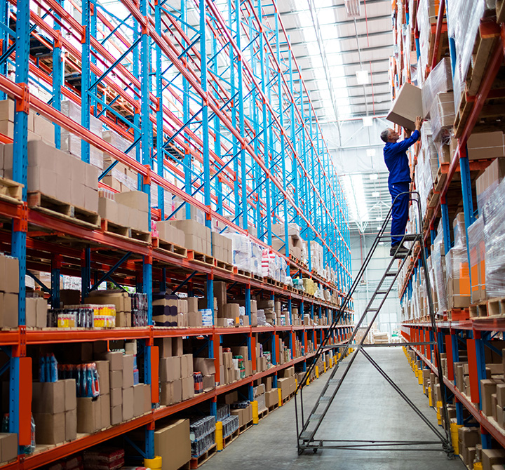 Man in warehouse on ladder placing a box onto a high shelf. 