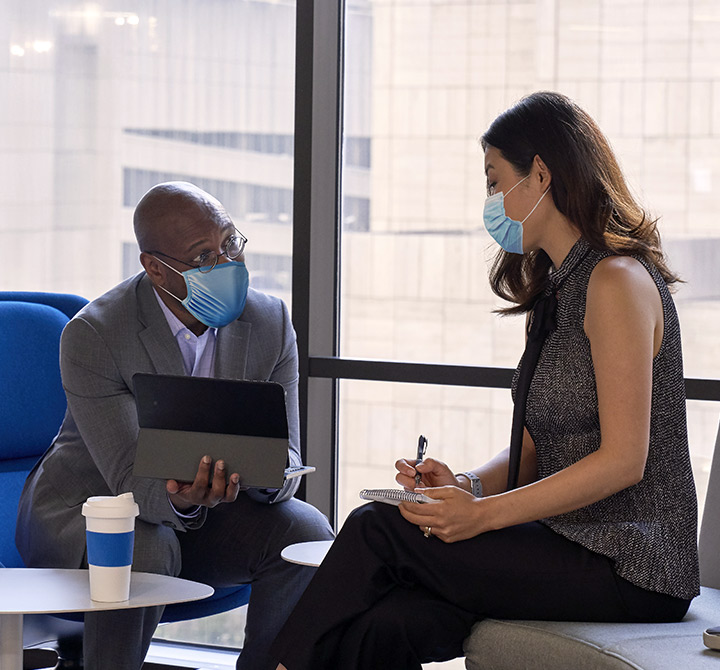 A man in glasses holding a tablet shows a woman he is meeting with something while she takes notes in a notebook.