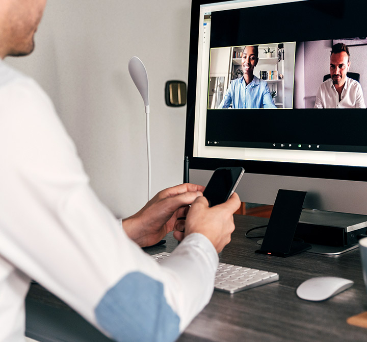 A man sitting at a desk in his home office is on a video conference call and holding a mobile phone in his hands.