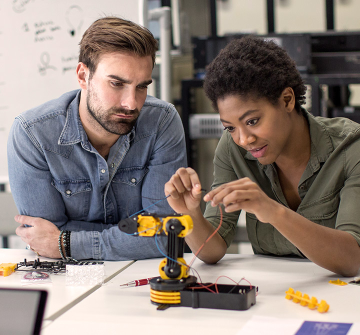 A male and female technology students are working together with the wires on an electrical device in a computer lab.