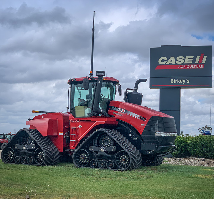 A large red tractor with large black tracks parked outside on the grass in front of a Birkey's road sign.