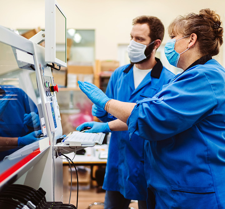 Two employees wearing blue smocks, medical gloves, and masks are reviewing a machines screen diagnostics.