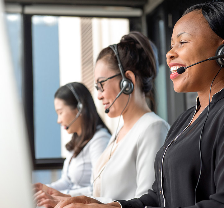 A female customer care rep, wearing a headset, is looking at a computer monitor, with colleagues in the background.