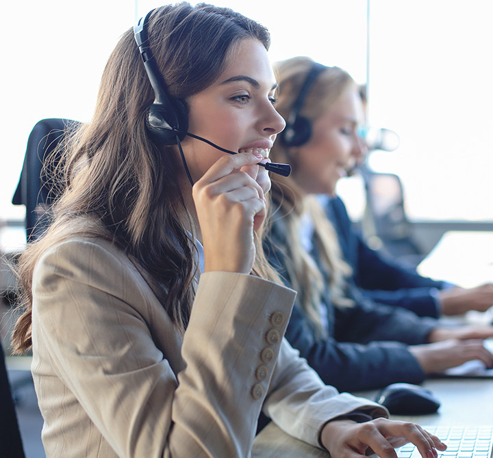 A female customer care rep, wearing a headset, is looking at a computer monitor, with colleagues in the background.
