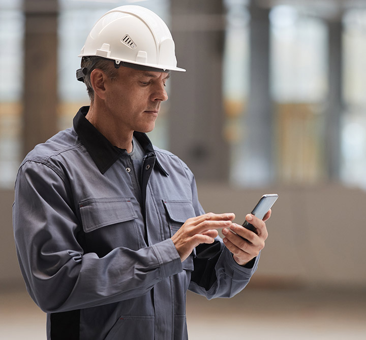 A male worker wearing a white hard hat and grey uniform is tapping on a mobile device he is holding in his left hand.