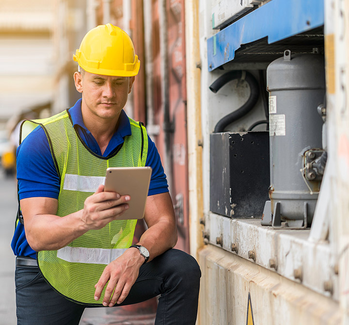 A male worker wearing a yellow hard hat and vest is working from a tablet device he is holding in his right hand.