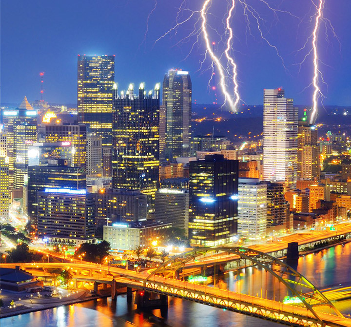 Cityscape at night time with bold, visible lightening strikes in the back ground.