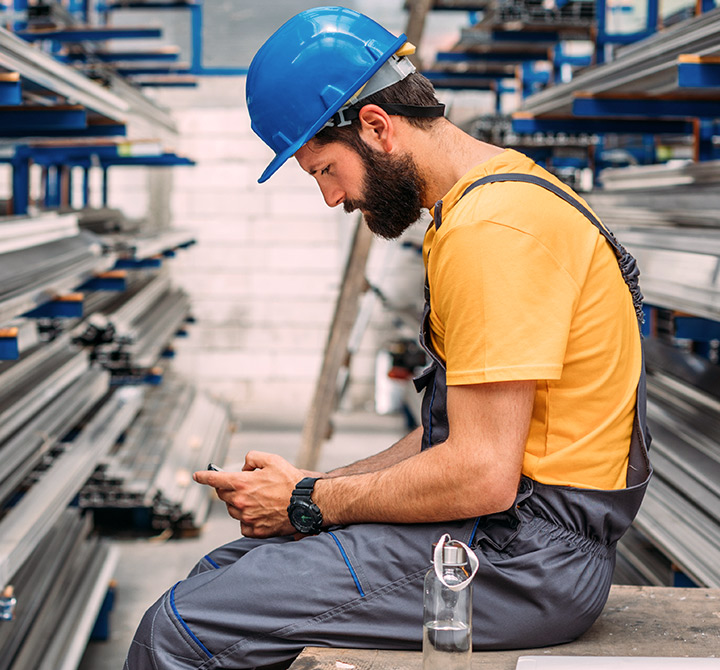 A man in gray coveralls, yellow shirt, and blue hard hat uses a mobile device.