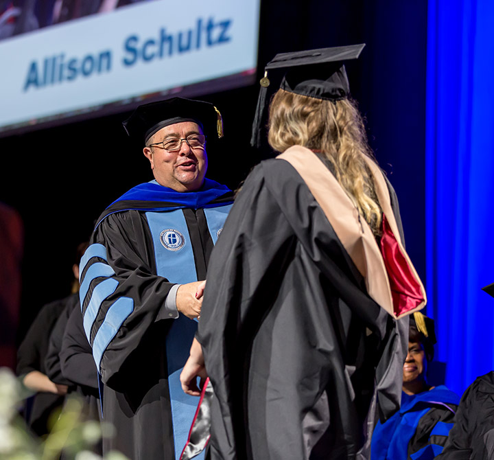 A view of a male college president shaking the hand of a graduating female student at a commencement ceremony.