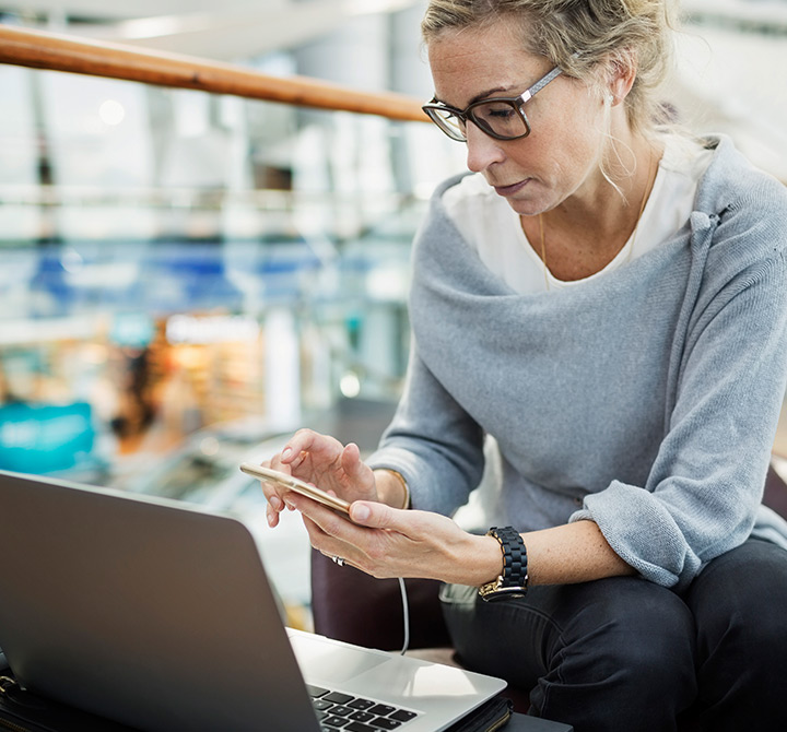 Businesswoman in a gray sweater looking at her phone while it is connected to her laptop charging.