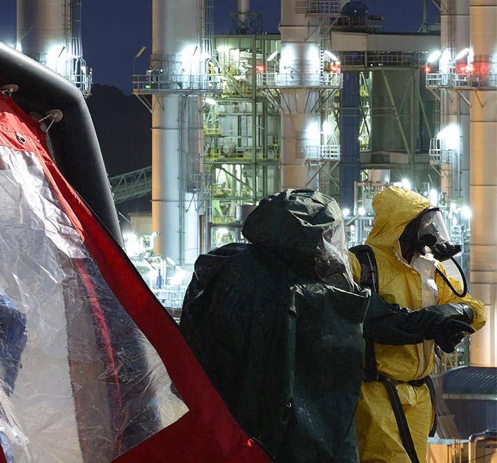 A worker in a black bio hazmat suit walks next to somebody in a yellow hazmat suit in an oil field setting.