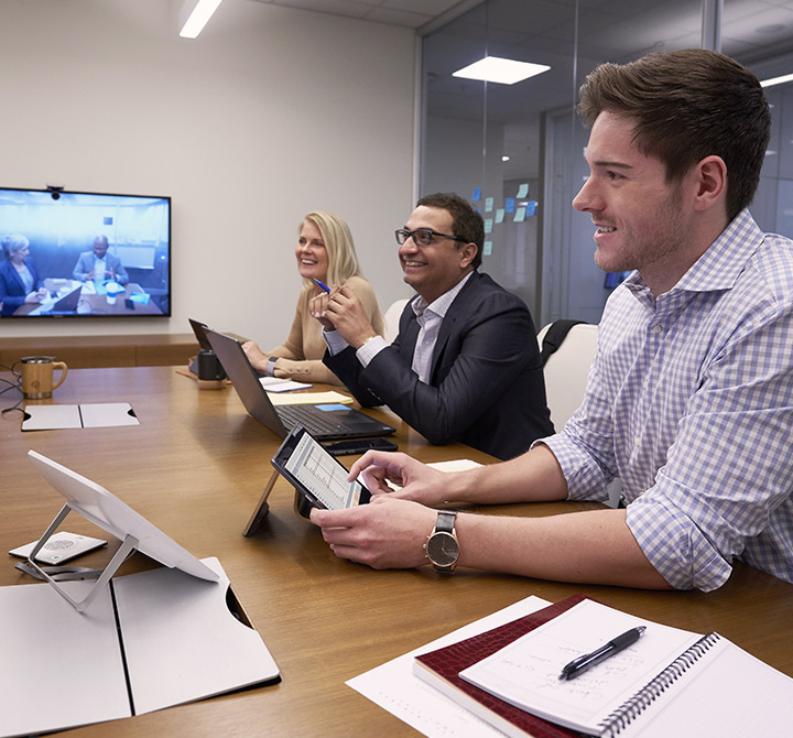 Man in a plaid, collared shirt is using a tablet in a large meeting with co-workers using live video chat.
