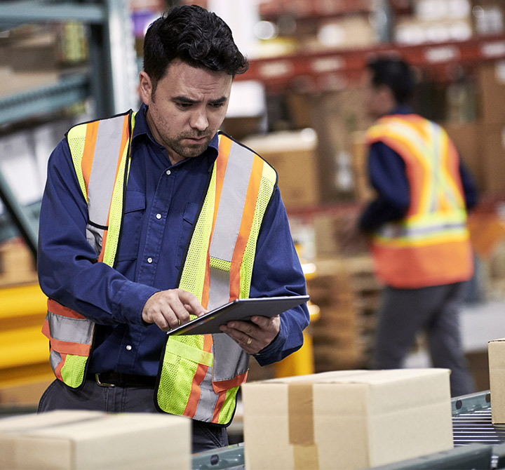A man in a blue shirt and yellow safety vests holds a tablet in a warehouse as packages go by on a conveyor belt.