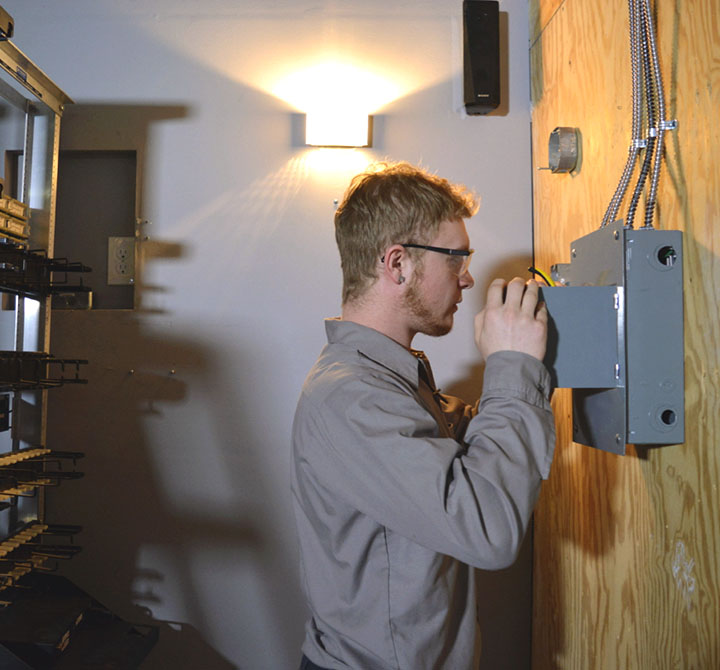 A male Lincoln Tech student, wearing safety glasses, is working on an electrical panel, as part of the education track.