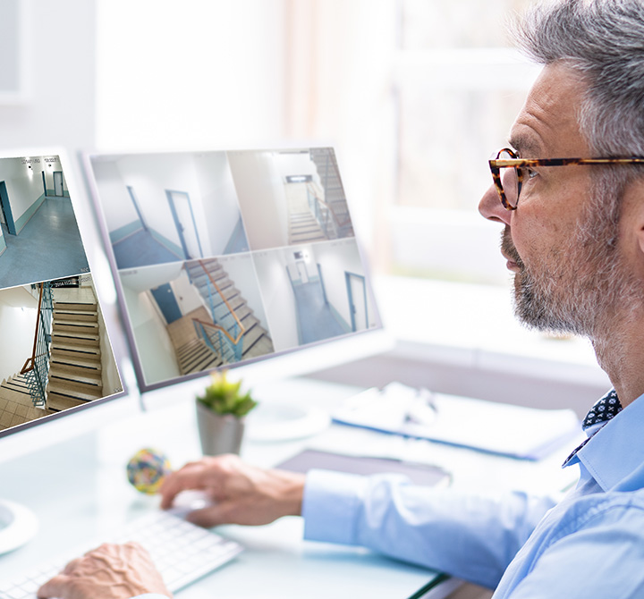 A man wearing a blue shirt and glasses is sitting at a desk in front of two computer monitors with a white keyboard.