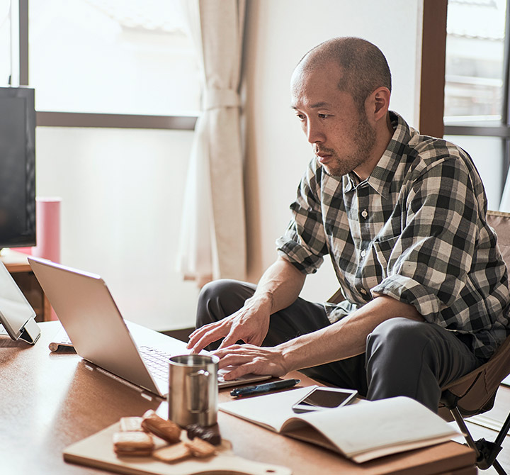 A man wearing a plaid shirt is sitting in front of a table typing on a laptop computer with his other mobile devices nearby.