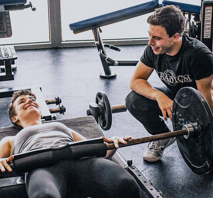 A male fitness trainer, wearing a black t-shirt and pants, is working with a client on an exercise machine in a gym.