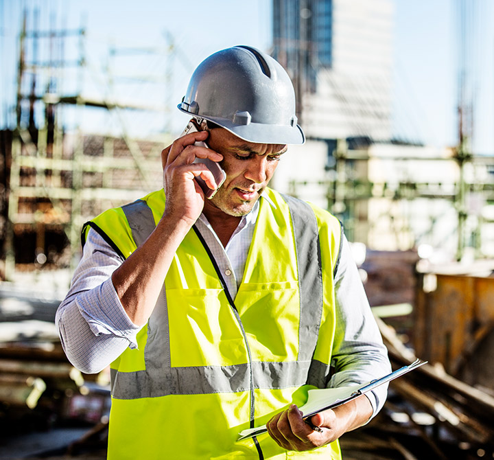 A male construction site worker wearing a hard hat and yellow safety vest is talking on his mobile device.