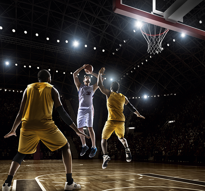 Image from a low angle viewpoint of a basketball player going to shoot a ball in a hoop basketball in front of rival players. The action takes place in generic indoor floodlit basketball arena full of spectators. All players are wearing generic unbranded basketball uniform.