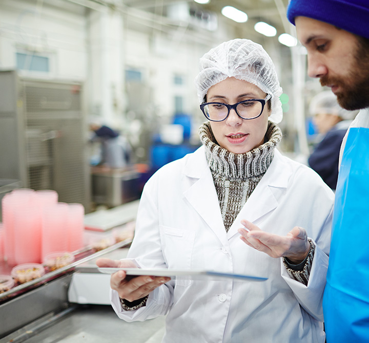 Woman in manufacturing plant holding a connected tablet while showing her coworker.
