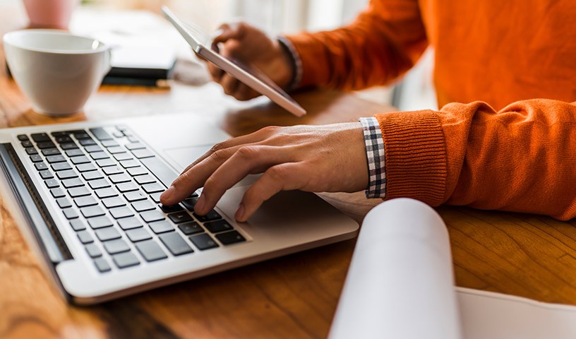 A man wearing a orange sweater, is sitting in front of a laptop computer looking down at his mobile device in his hand.