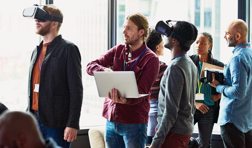Two mean with Oculus headsets on look around in VR as another man in a red shirt holding a laptop goes through a presentation with a diverse group talking in the background.