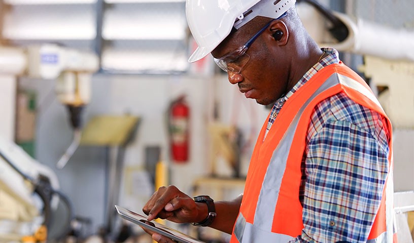 A man in a white safety helmet and orange safety vest is using a tablet in a factory.