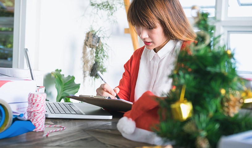 A woman in a white shirt and red cardigan is writing on a clipboard with Christmas decorations in the foreground.