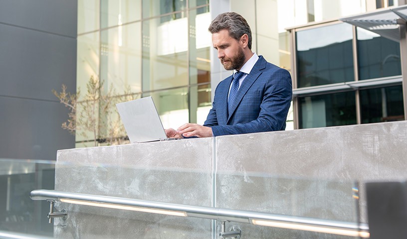 A man in a blue suit uses his laptop outside of his office building.