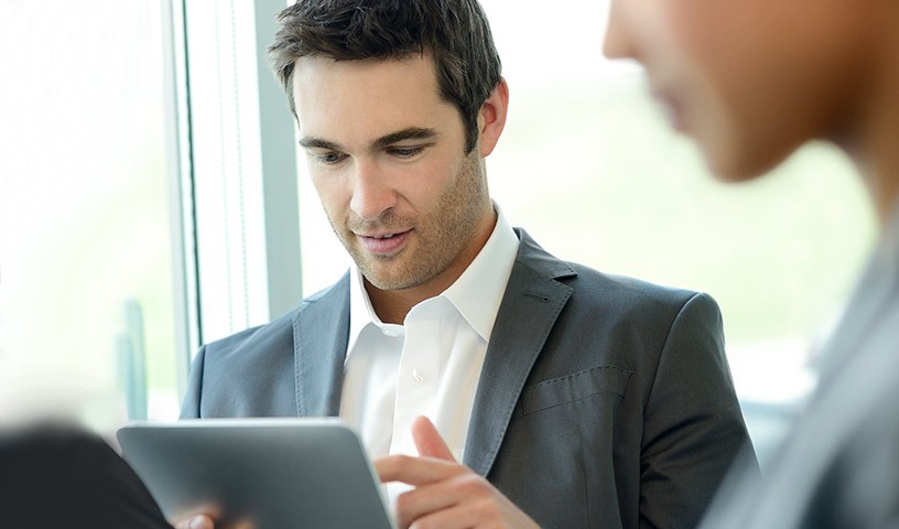 A man wearing a grey suit is sitting in a conference room with others doing work from his tablet device.