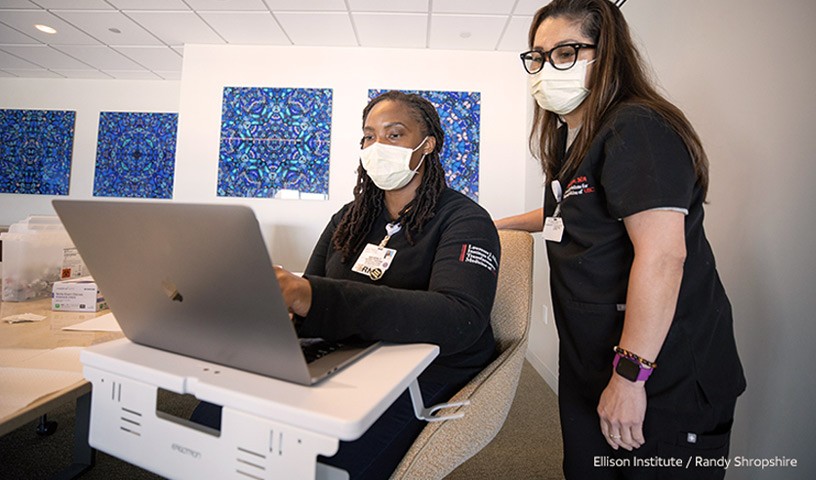 Two medical technicians wearing uniforms and masks are looking at a laptop computer to perform their job. 