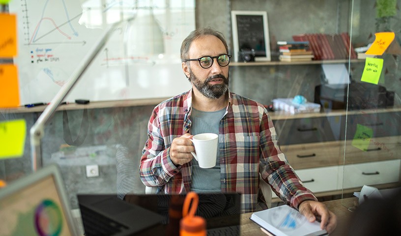 A bearded man wearing a plaid shirt and glasses is sitting at a desk, looking at a computer monitor holding a cup of coffee.