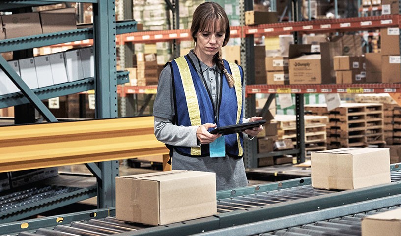 A woman wearing a blue safety vest with yellow stripes in a warehouse holds a tablet device while surveying boxes moving along a conveyor belt.