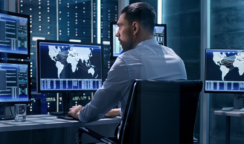 A man sits at a desk monitoring the networks security while in front of multiple monitors.