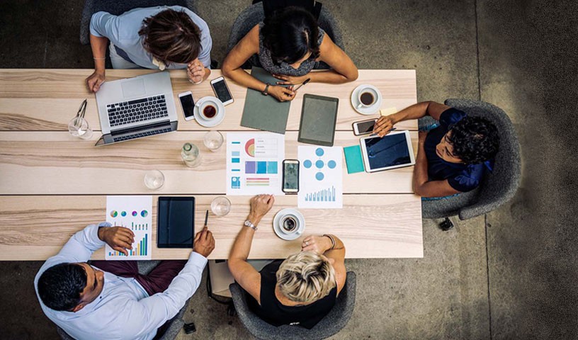 A group sits a table having a meeting with print outs and their laptops on the table.
