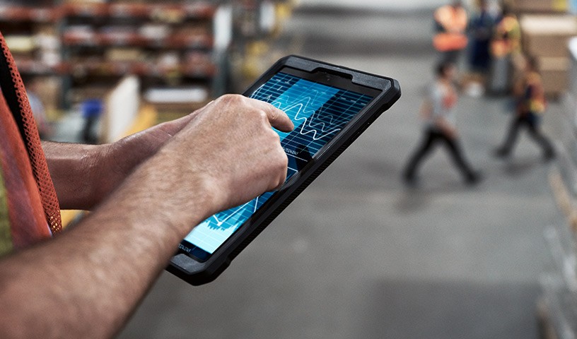 Man in a warehouse setting looking at a tablet while workers work in the background.