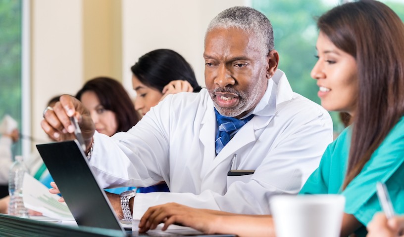 Senior adult African American doctor and professor is assisting a young adult Hispanic female nursing or medical student in class. Doctor is pointing to laptop computer screen while nursing student types. Other healthcare students are studying around them.