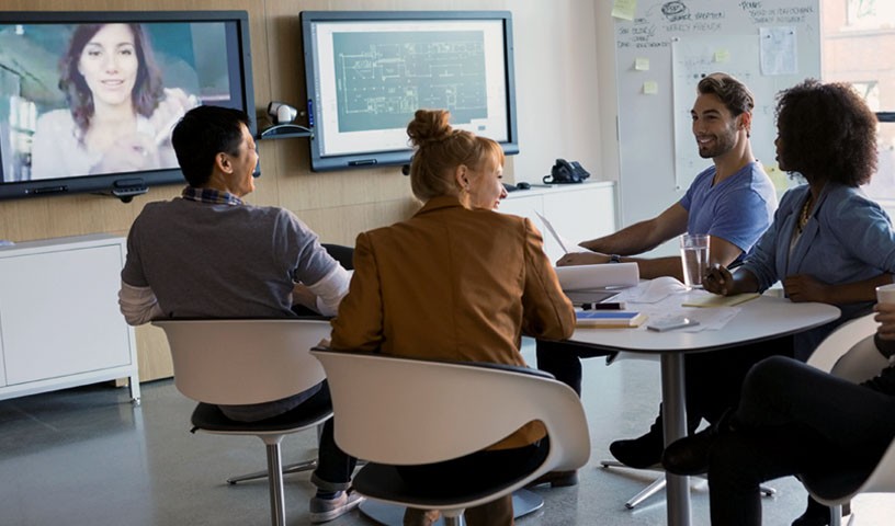 A group of people are sitting in a meeting room on a video call with a 5th member.