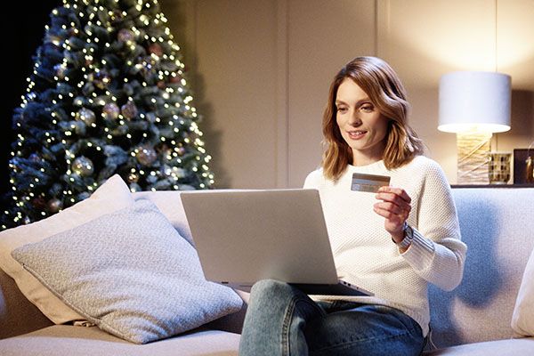 Cheerful smiling beautiful woman buying on internet xmas presents paying with credit card using laptop at cozy decorated home. Xmas sale and discounts. Holiday shopping