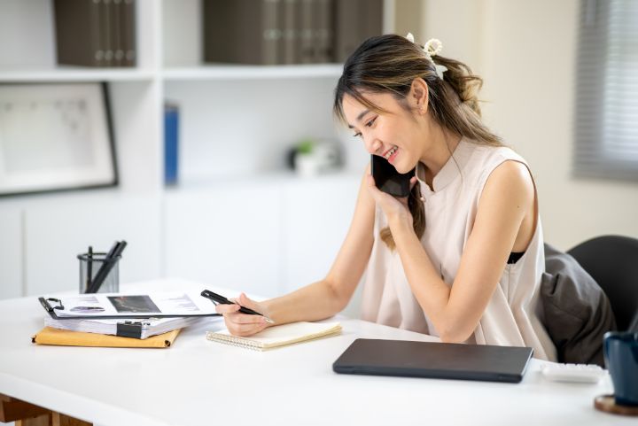 Pretty woman office worker holding pen over notebook while talking on phone at white table in office. Business Casual, Modern Company, Corporate Job.