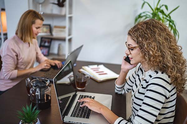 Two female colleagues work together at home using laptops while one of them makes a phone call