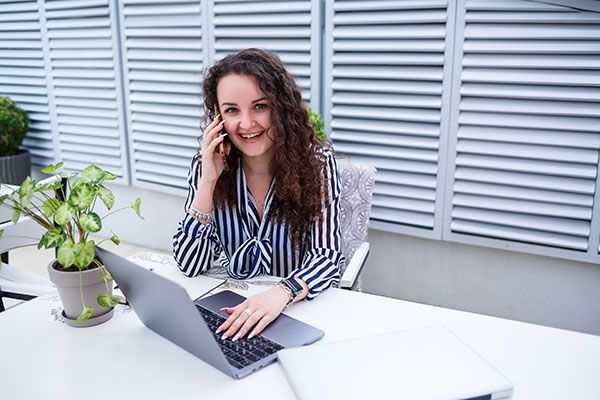 Smiling successful young woman talking on the phone, working on a laptop on the terrace, girl in a cafe at a business meeting. Business concept