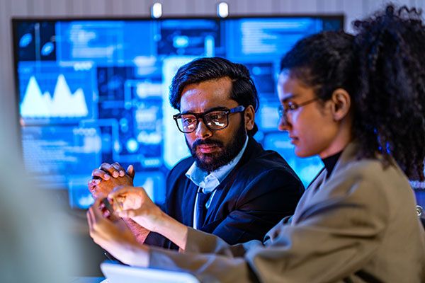 South Asian male and Black female cybersecurity professionals review digital forensics data in a blue-lit dark operations center with multiple security screens active.